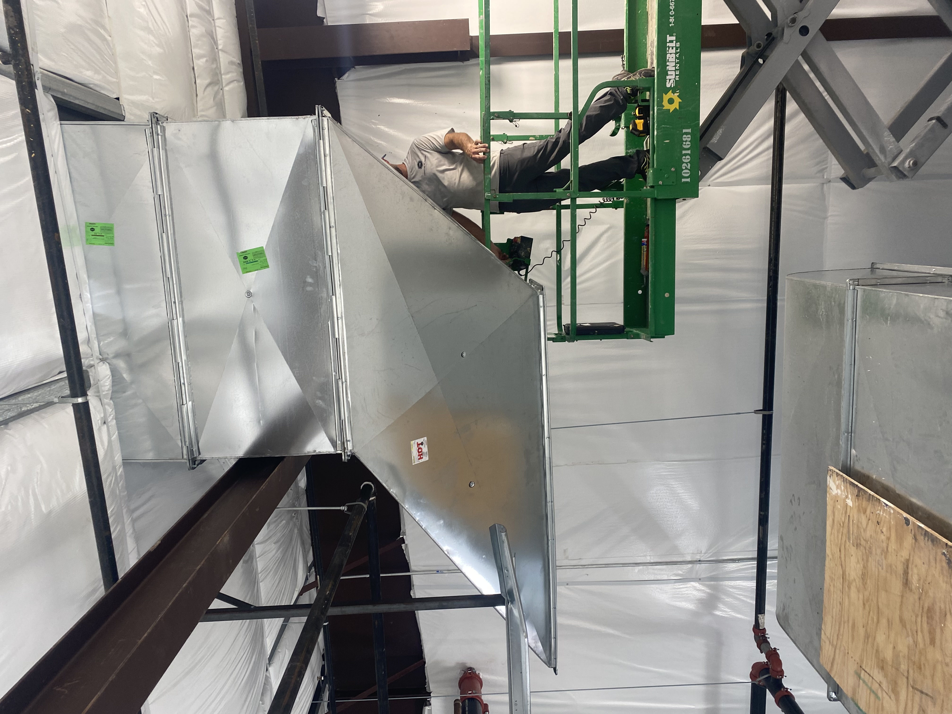 CFM technician installing large rectangular ductwork from a scissor lift at an auto repair center